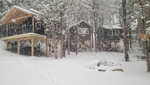 Une cabane en bois dotée d’un porche couvert et une plus petite cabane, le tout situé dans une forêt enneigée.
