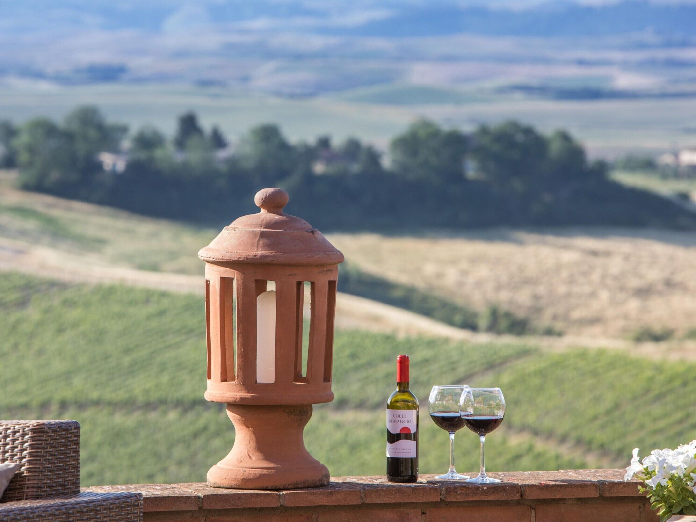 A terracotta lantern, a bottle of wine, and two wine glasses on a brick ledge with a view of rolling hills.