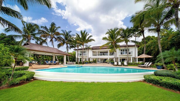 Ein großer Swimmingpool, umgeben von üppiger Vegetation und Palmen, mit einem mehrstöckigen Gebäude im Hintergrund.