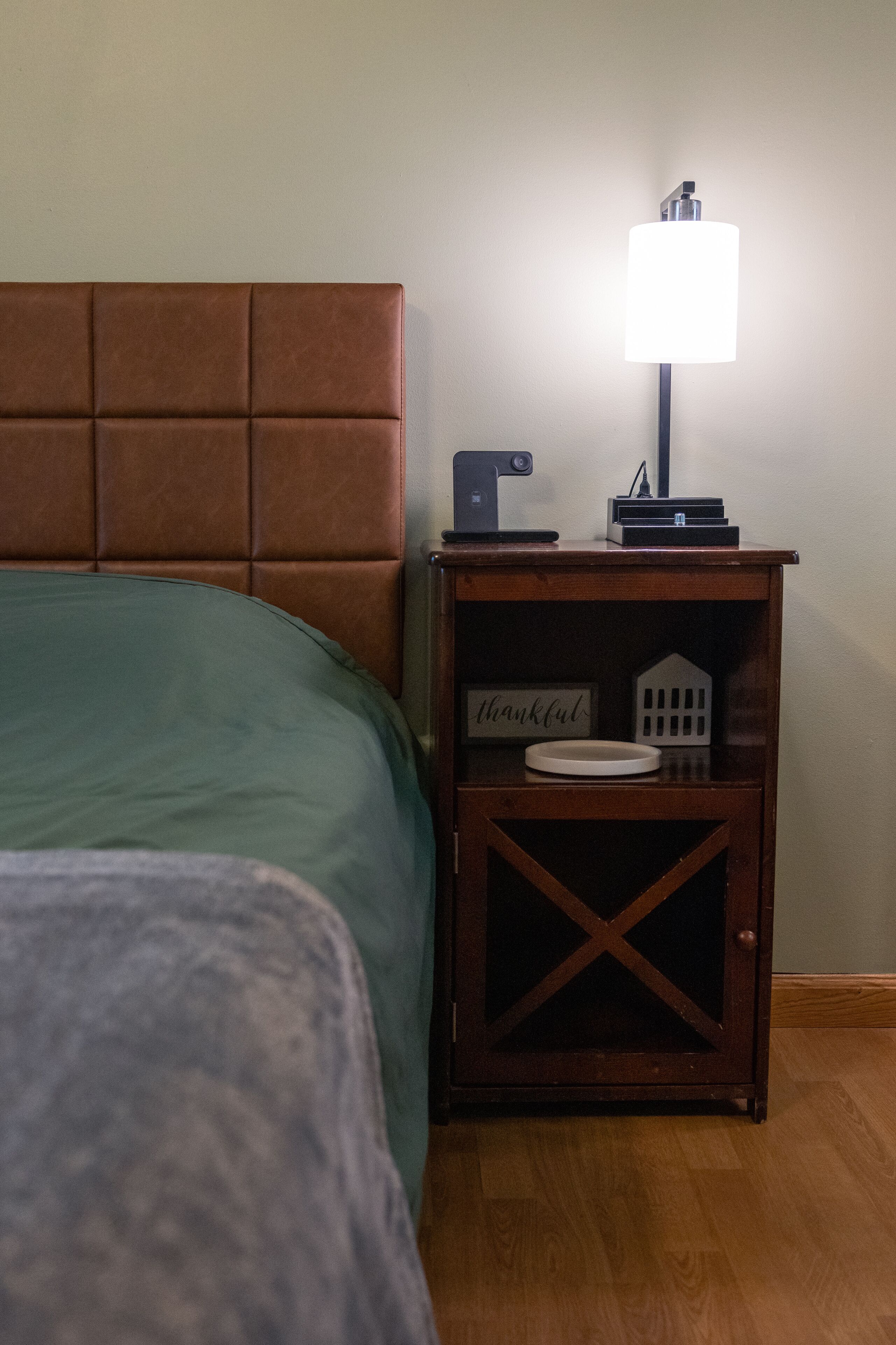 A neatly made bed with a green bedspread and a wooden headboard. A small wooden nightstand with a lamp and a sign that reads "thankful" is beside the bed.