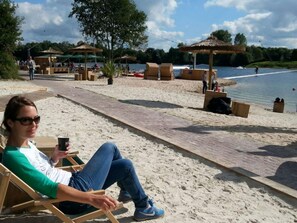 Una persona sentada en una silla de playa sosteniendo una bebida, con un paseo marítimo de madera y sombrillas de playa al fondo.