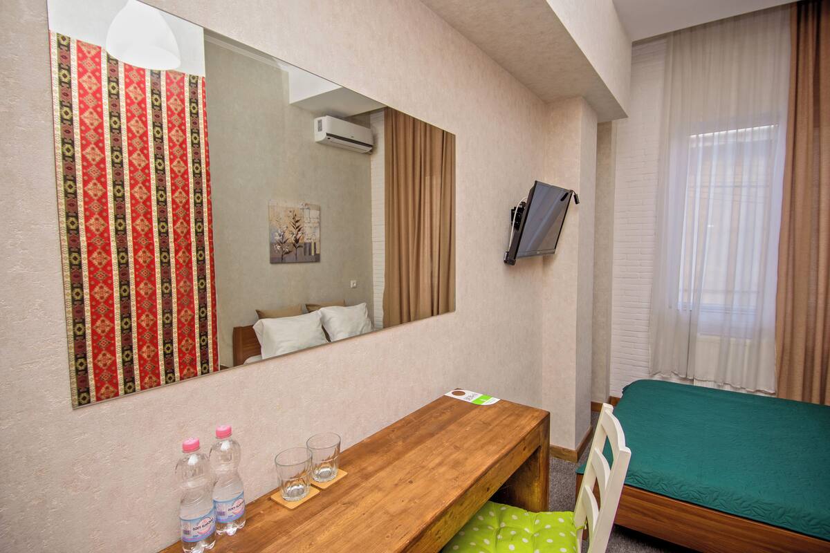 Een hotelkamer met een houten bureau, een bed met een groene spreidek, een wandtelevisie, een spiegel en een raam met gordijnen.