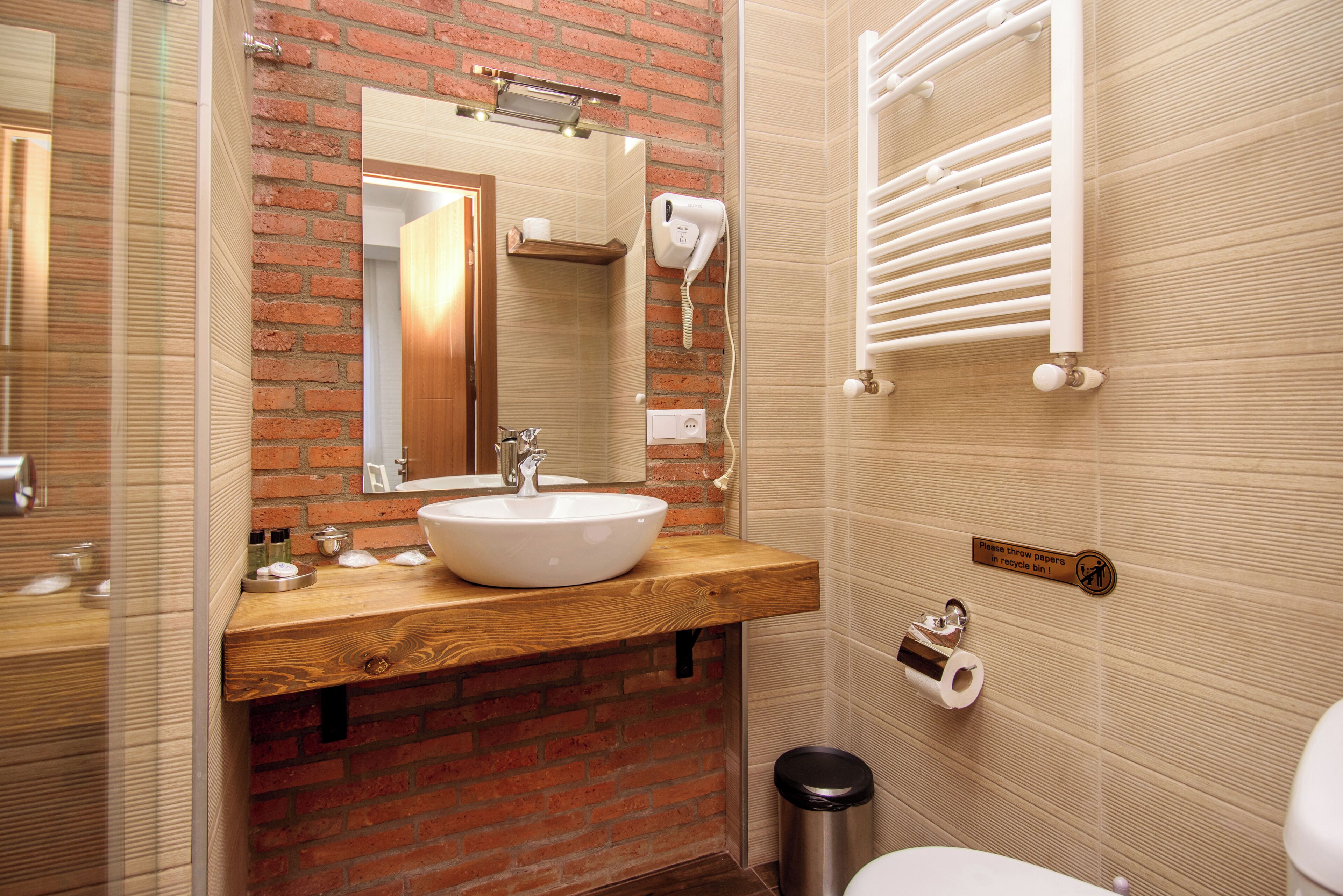 Un baño con tocador de madera, un lavabo blanco, un espejo, un secador de pelo, toallero y un contenedor de reciclaje.