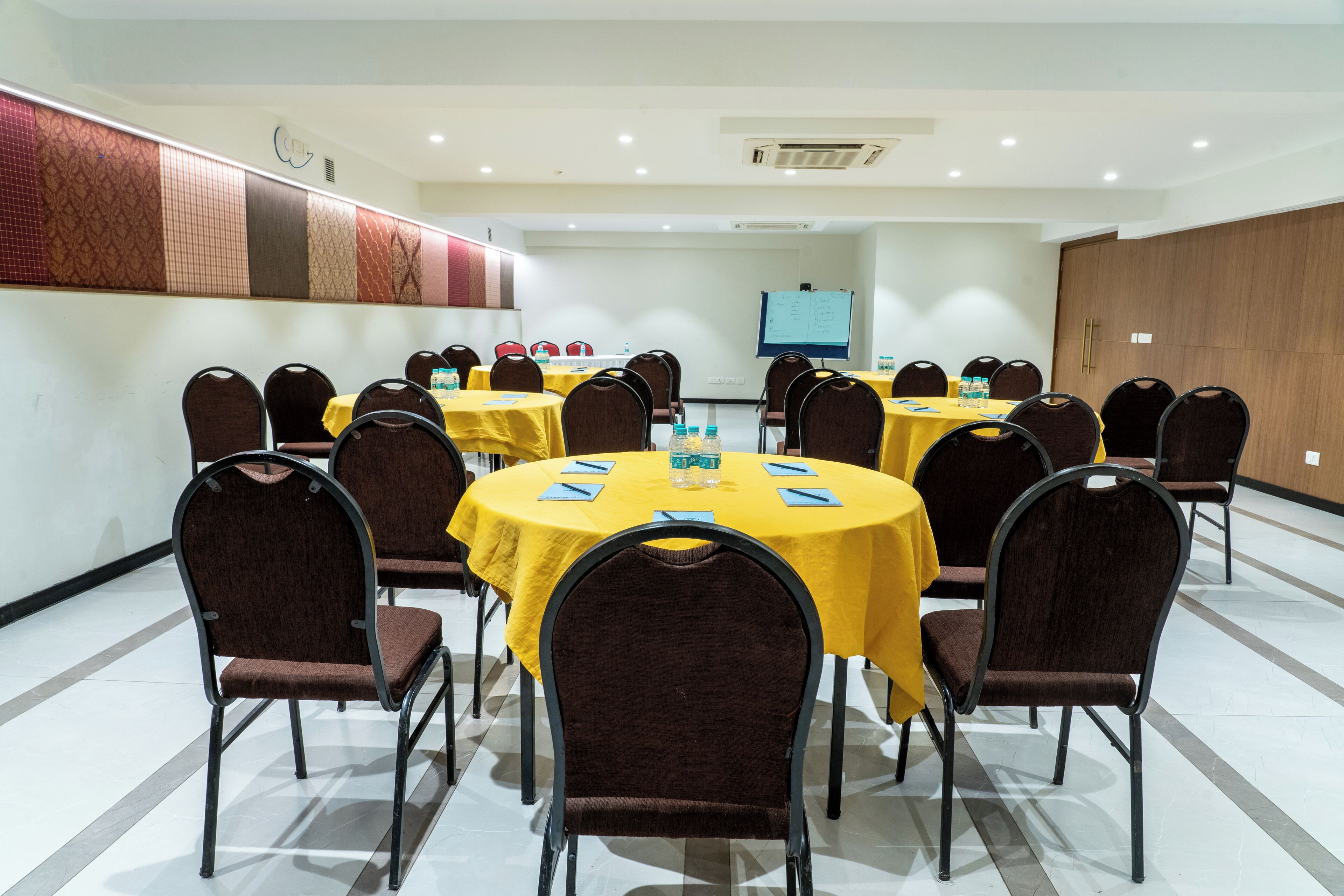 Sala de conferência com várias mesas e cadeiras, equipada com garrafas de água e cadernos.