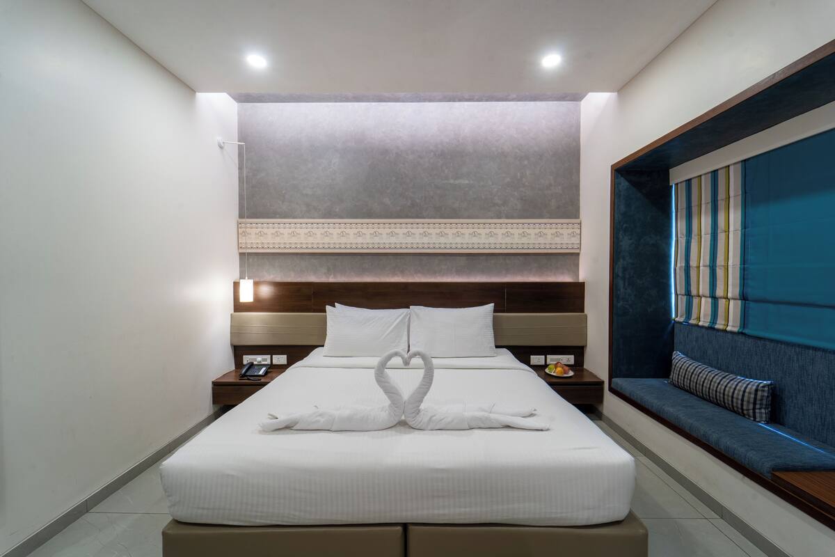 Habitación de hotel con una cama bien hecha, una mesita de noche con un teléfono, una mesita de noche con una lámpara y un banco empotrado con cojín.