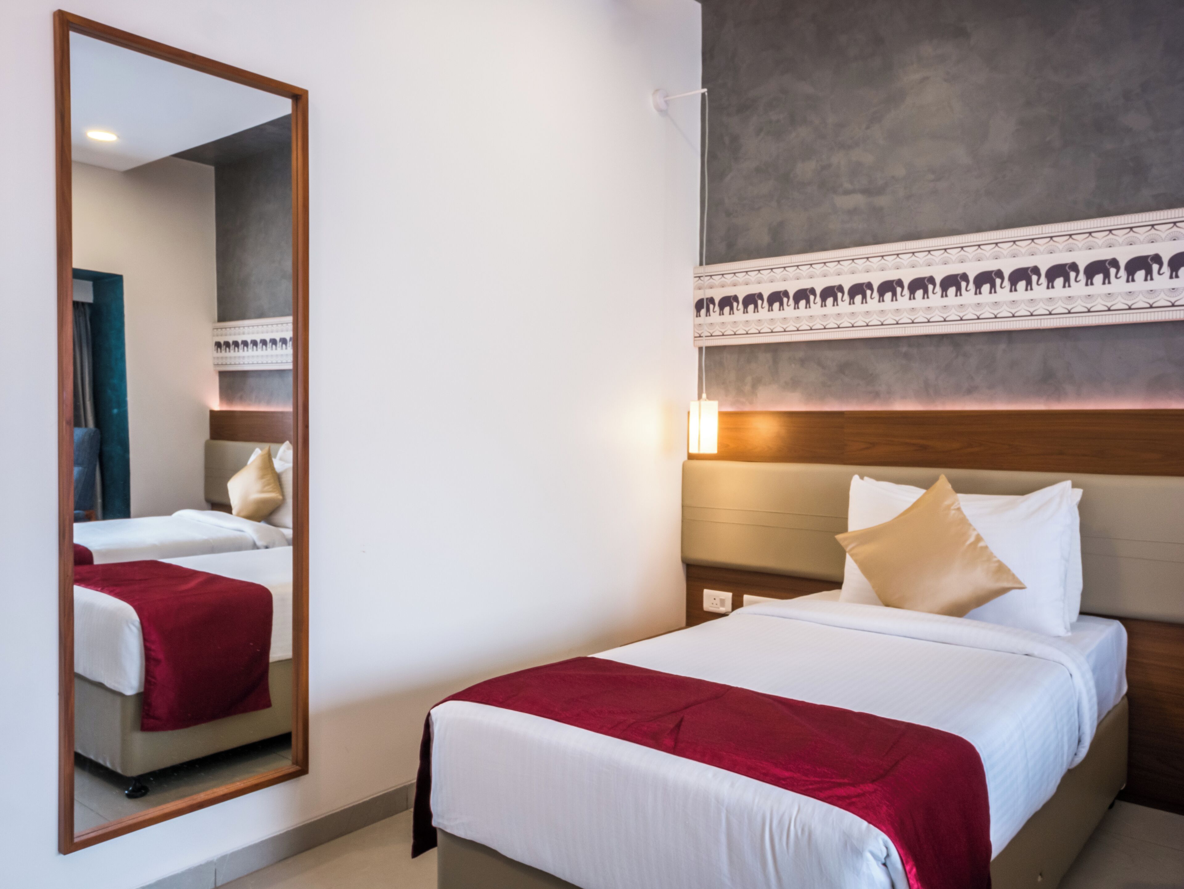 Habitación de hotel con un espejo grande, una cama con ropa de cama blanca y roja, un cabecero con un diseño decorativo y una mesita de noche montada en la pared con una lámpara.