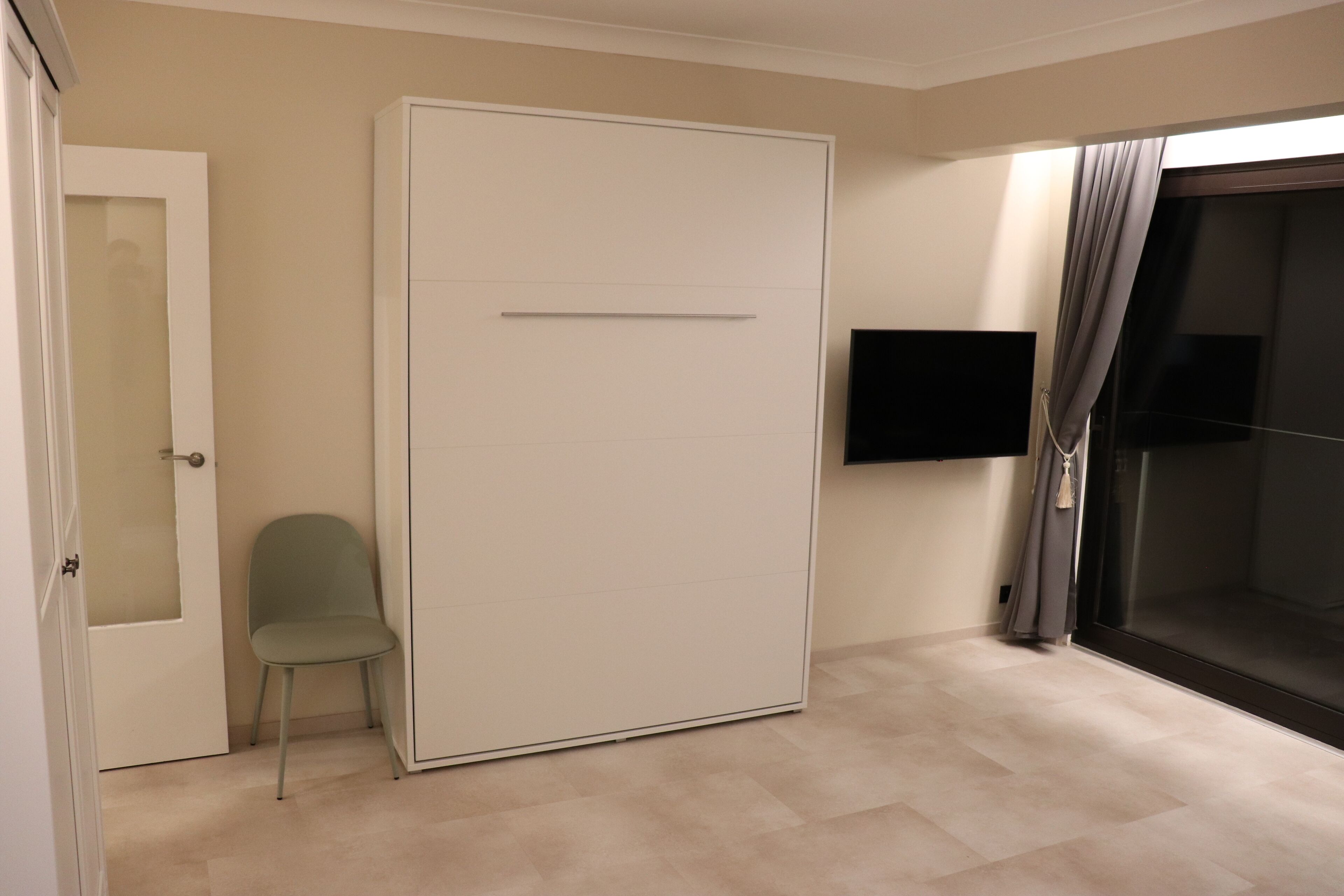 Quarto com porta deslizante, uma cadeira, TV de tela plana na parede e uma janela grande com cortinas.