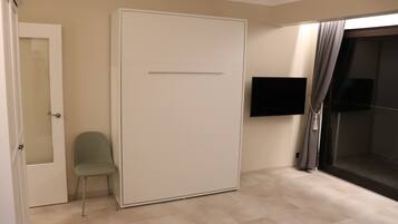 Quarto com porta deslizante, uma cadeira, TV de tela plana na parede e uma janela grande com cortinas.