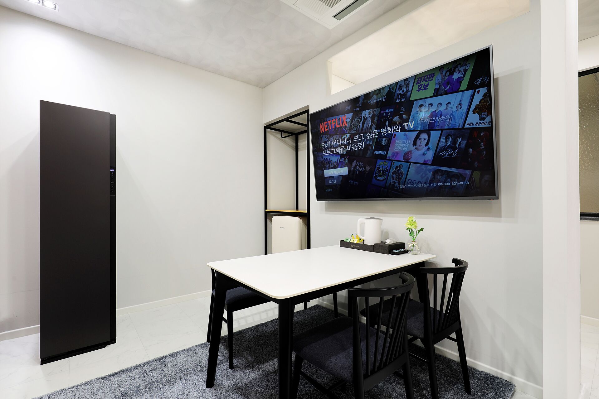 Una habitación moderna con una mesa blanca y sillas negras, un televisor grande en la pared mostrando diverso contenido, y un armario negro.
