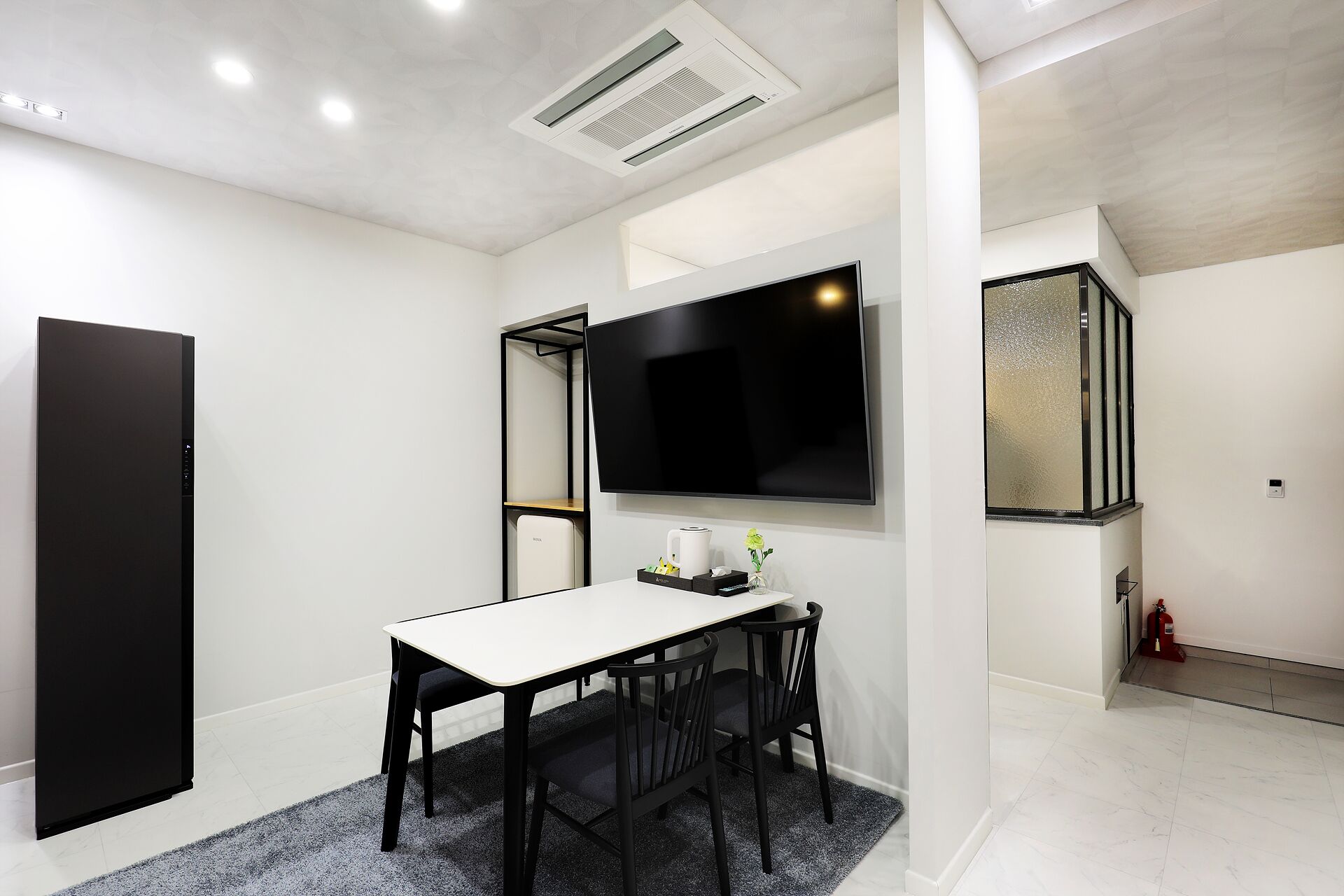 A modern dining area with a white table and black chairs, a flat-screen TV mounted on the wall, and a small side table with a vase and a coffee maker.