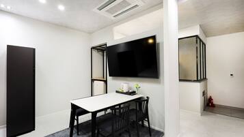 Un moderno comedor con una mesa blanca y sillas negras, un televisor de pantalla plana montado en la pared y una mesita auxiliar con un florero y una cafetera.