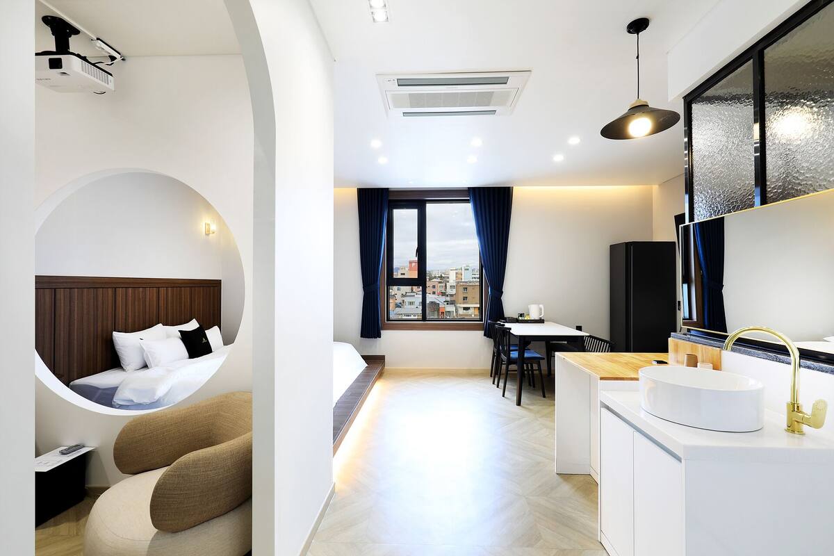A modern hotel room with a bed, a circular seating area, a dining table, and a kitchen area.