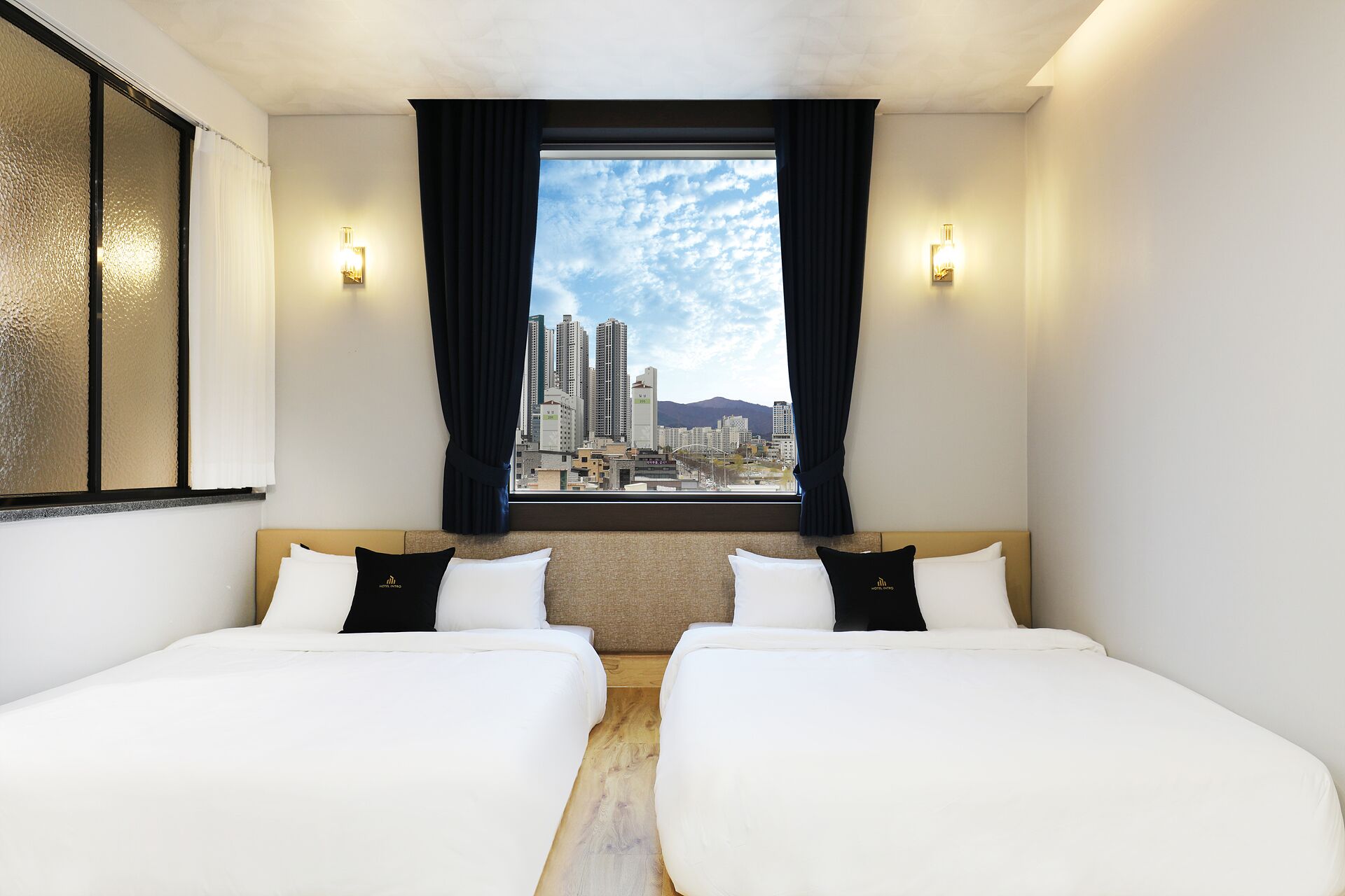 Habitación de hotel con dos camas, vista a la ciudad y dos luces fijadas a la pared.