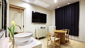 A hotel room with a white sink, a mirror, a flat-screen TV mounted on the wall, a small table with two chairs, and a window with dark curtains.