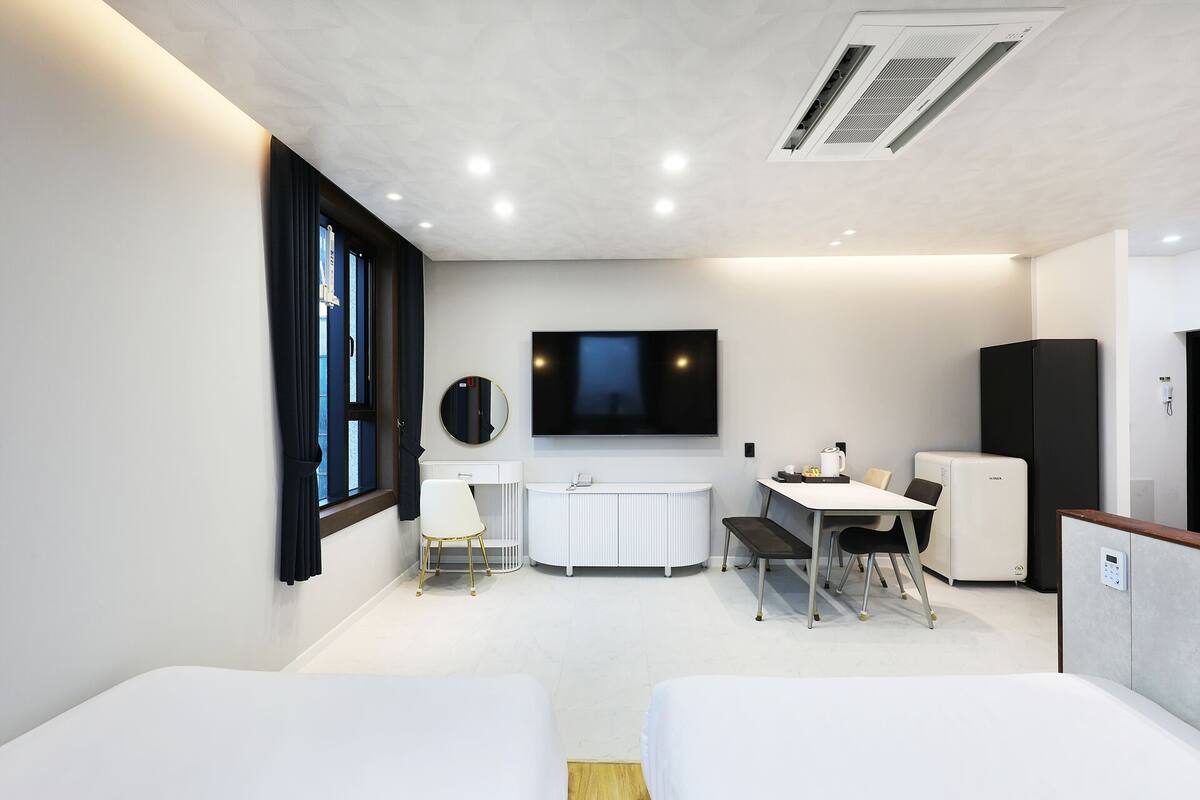 A modern hotel room with two beds, a TV, a desk with a chair, a refrigerator, and a bathroom.