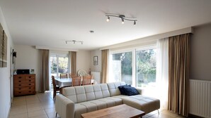 A spacious living room with a beige sofa, a wooden coffee table, and a dining area with a table and chairs. - Private Garden Near Sea (De Haan)