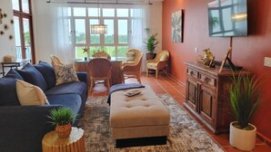 A living room with a sofa, ottoman, and chairs, a dining area with a table and chairs, a TV mounted on the wall, and a large window with a view of greenery.