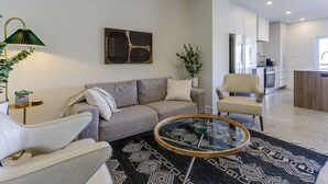 Ein modernes Wohnzimmer mit einer Couch, einem Sessel und einem Couchtisch, im Hintergrund eine Küche und ein großes abstraktes Gemälde an der Wand.