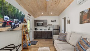 Un ambiente compatto con angolo cottura, un divano con cuscini a righe, soffitto in legno e vista su veicoli fuoristrada attraverso un'ampia finestra.
