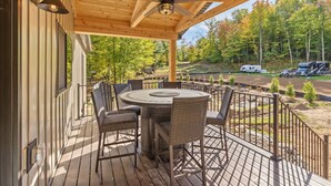 Un patio in legno con un tavolo rotondo e sedie in vimini, con vista su una zona boschiva che include una strada e camper.