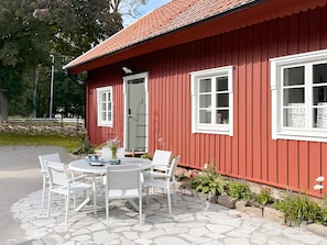 A red wooden building with two white-framed windows and a door, a stone patio with a round table and four chairs, and a small garden area with plants and a bird feeder. - 4 Star Holiday Home in Tvaaker (Tvaaker)