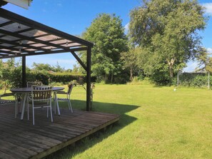 A wooden deck with a table and chairs, a covered area with a roof, and a grassy field with trees and a soccer goal. - 6 Person Holiday Home in Stubbekobing (Stubbekobing)