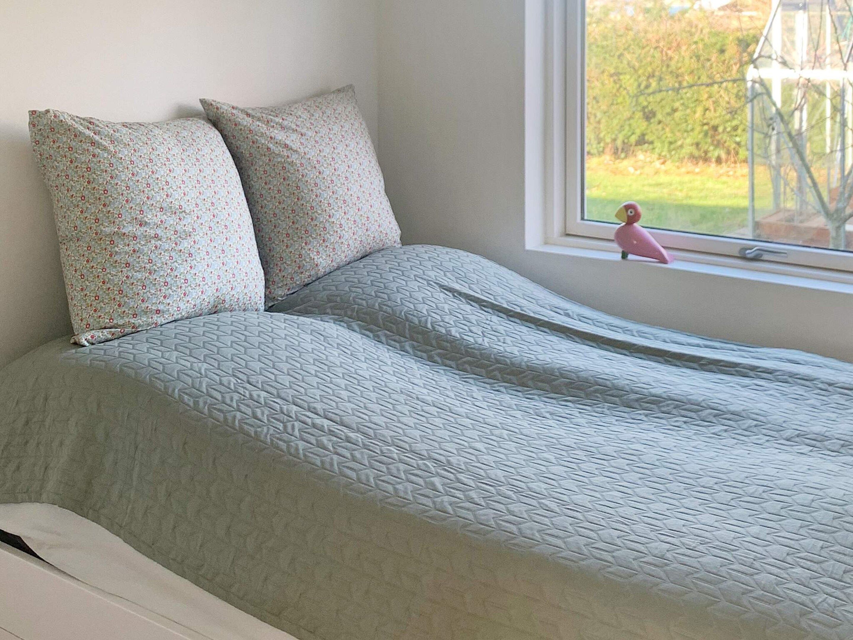 Un lit avec une couvre-lit à motifs et des oreillers, une fenêtre donnant sur un paysage verdoyant, et un petit jouet rose posé sur le rebord de la fenêtre.