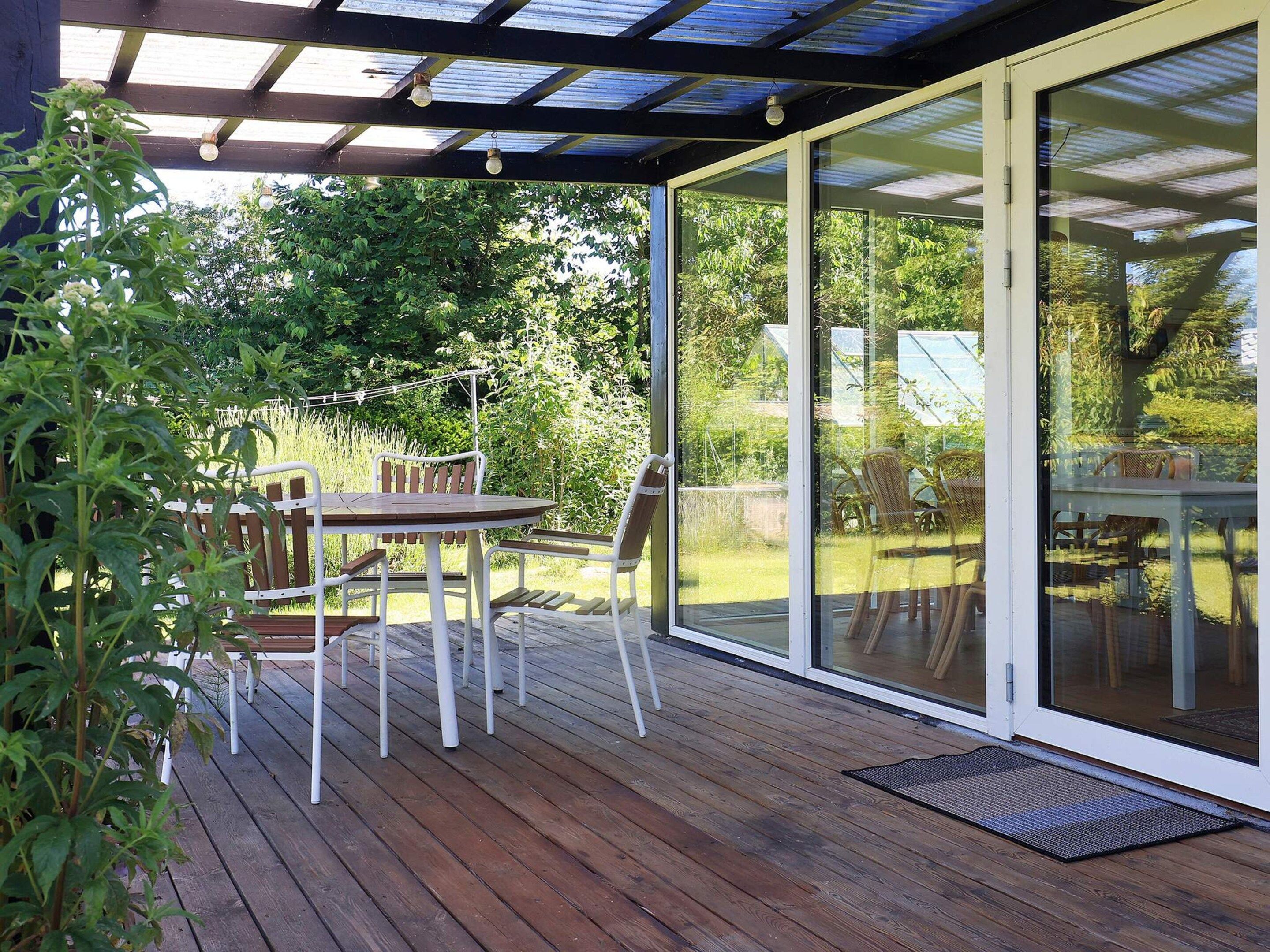 Eine überdachte Terrasse mit Tisch und Stühlen, ein Holzdeck und eine Glastür zum Garten.