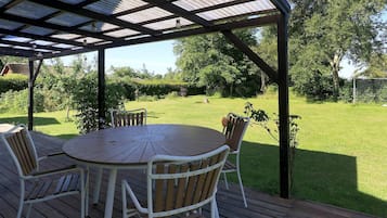 An outdoor patio with a round table and chairs, a metal roof, and a view of a grassy area with trees.