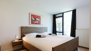Un dormitorio moderno con una cama de madera, una mesita de noche, una lámpara de noche y una ventana con vistas a árboles.