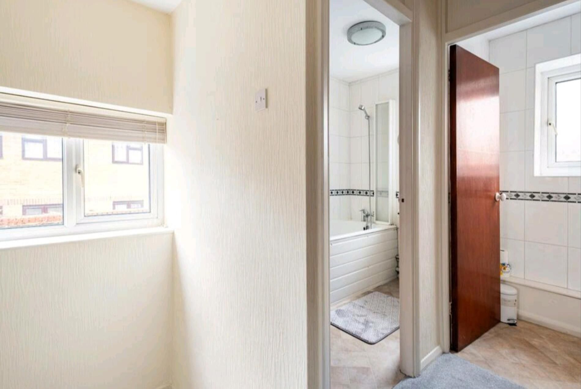A bathroom with a shower, a toilet, and a sink. There is a window with blinds and a door leading to another room.