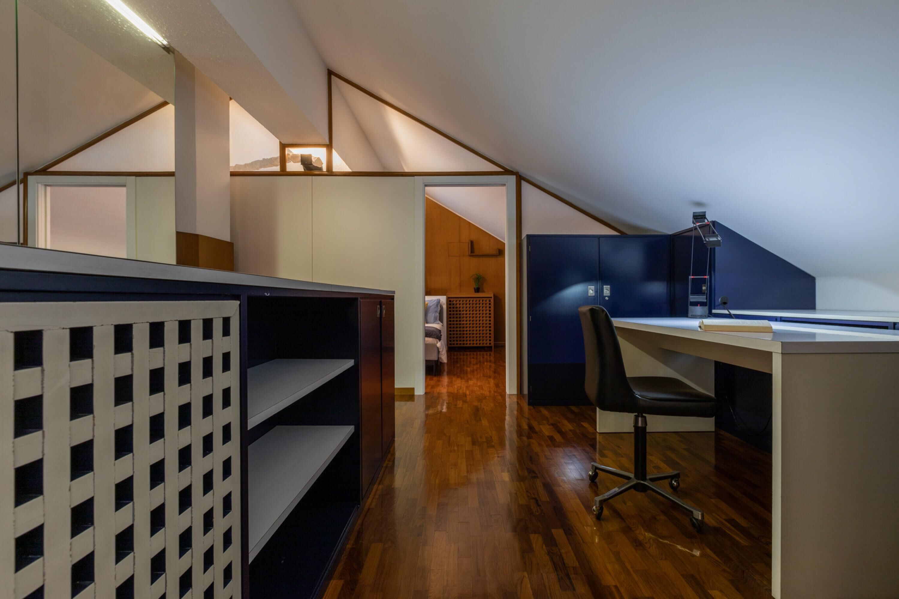 Quarto moderno estilo loft com teto inclinado, estantes embutidas, escrivaninha e cadeira.