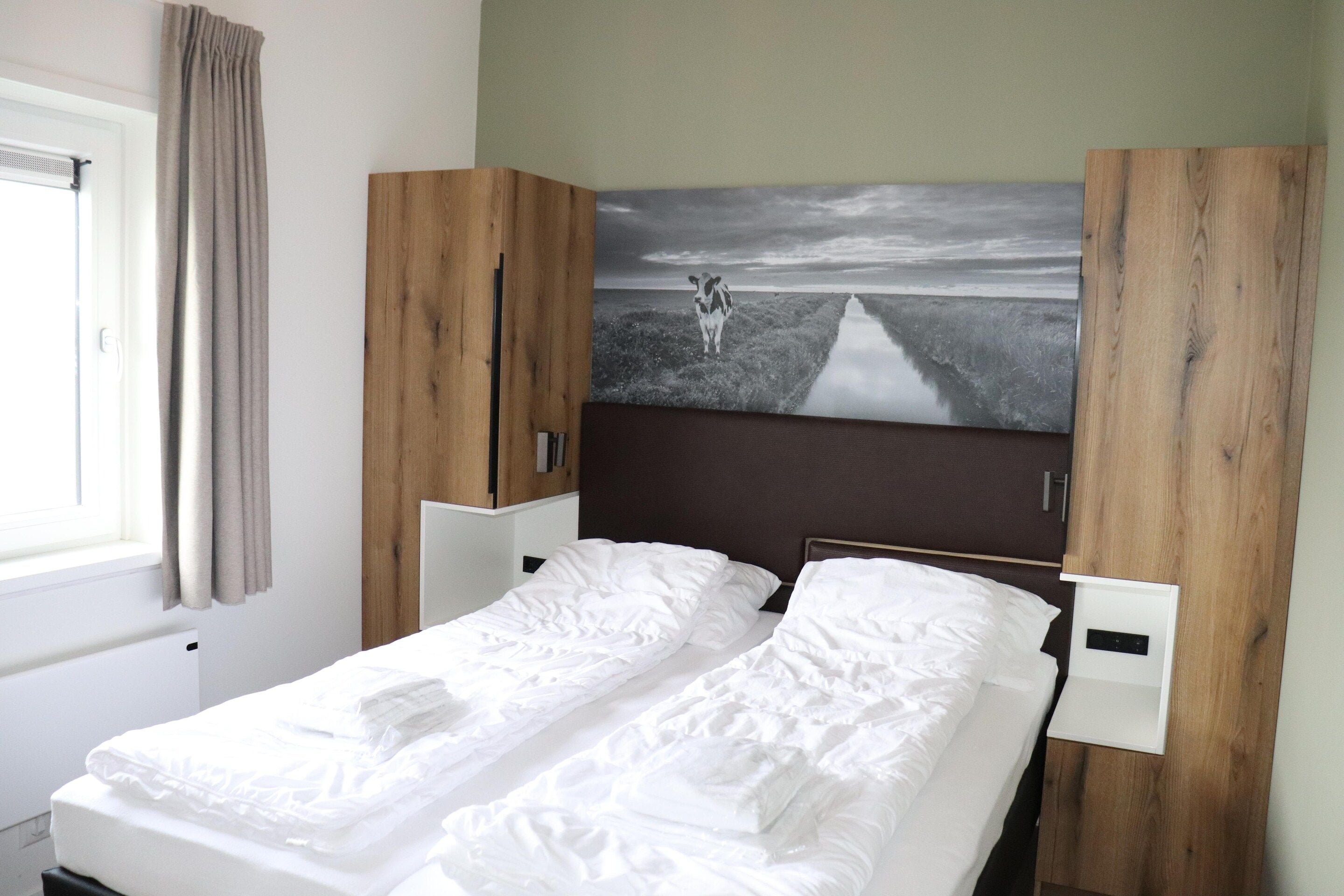 A single bed with white bedding, a wooden headboard, a wall-mounted light, and a painting of a landscape.
