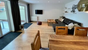 A modern living room with a sofa, wooden chairs, a coffee table, and a TV mounted on the wall. - Nice Apartment in Hopfen am See (Fuessen)