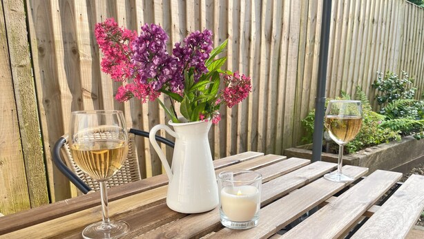 Ett träbord med två glas vitt vin, en vit kanna med lila blommor och ett tänt ljus.