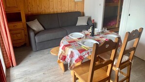 A cozy room with a wooden floor, a grey sofa, a dining table with a quilted tablecloth, wooden chairs, and a bunk bed in the background.