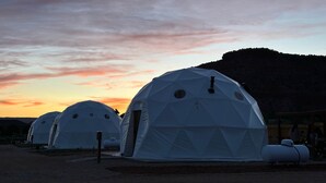 Des tentes en forme de dôme géodésique, dotées d’un toit plat, sont disposées en rangée, se détachant sur le fond d’un coucher de soleil, avec une colline visible au loin.