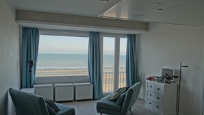 A modern room with a large window overlooking a beach, two teal armchairs, a white dresser, and a floor lamp. - Apartment in Nieuwpoort With Stunning Sea View (Nieuwpoort)