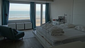 A bedroom with a large window overlooking a beach, a bed with white linens, a bedside table with a lamp, and a chair. - Apartment in Nieuwpoort With Stunning Sea View (Nieuwpoort)
