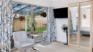 Une chambre moderne dotée d’une porte-fenêtre coulissante donnant sur un jardin, d’un fauteuil gris, d’une petite table, d’un ventilateur et d’un lit avec une tête de lit en bois.