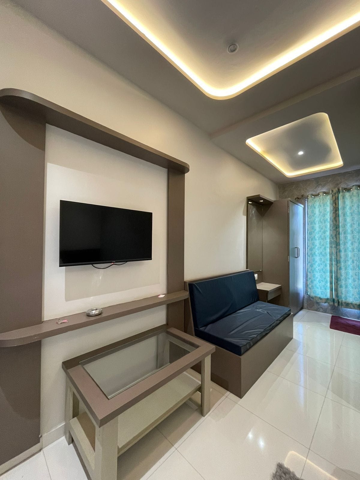 Una habitación de hotel moderna con una televisión de pantalla plana montada en la pared, una mesa de centro de vidrio, un sofá y una zona de ducha con cortinas.