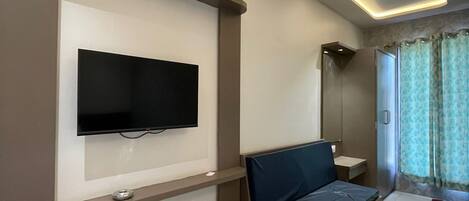 Una habitación de hotel moderna con una televisión de pantalla plana montada en la pared, una mesa de centro de vidrio, un sofá y una zona de ducha con cortinas.