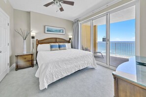 A bedroom with a large bed, a nightstand, a bedside table, a glass desk, and a view of the ocean through a sliding glass door. - Emerald Isle Resort 1504 (Panama City Beach)