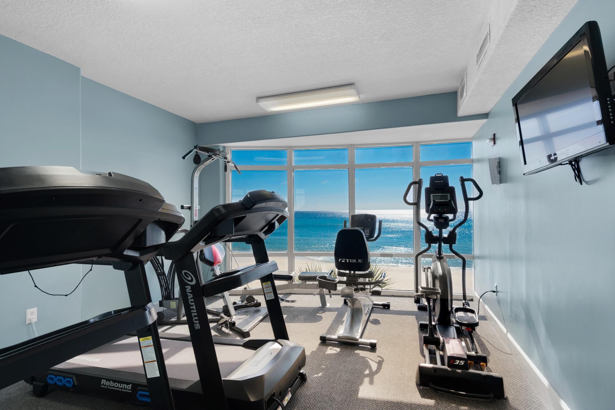 A gym with Nautilus and Rebound equipment, large windows with a view of the ocean, and a flat-screen TV on the wall.