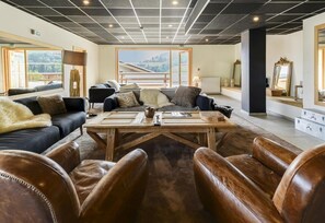 A spacious lounge area with leather seating, a wooden coffee table, and a large window offering a view of the outdoors. - Wifi, 200m from resort center, swimming pool, sauna, hammam, terrace, balcony, tv, ski locker (Combloux)