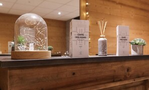 Un comptoir d’accueil en bois surmonté d’un dôme de verre contenant une plante, deux boîtes étiquetées « CARTE DES SOINS » (soins du corps) et un vase décoratif avec des tiges de roseau.