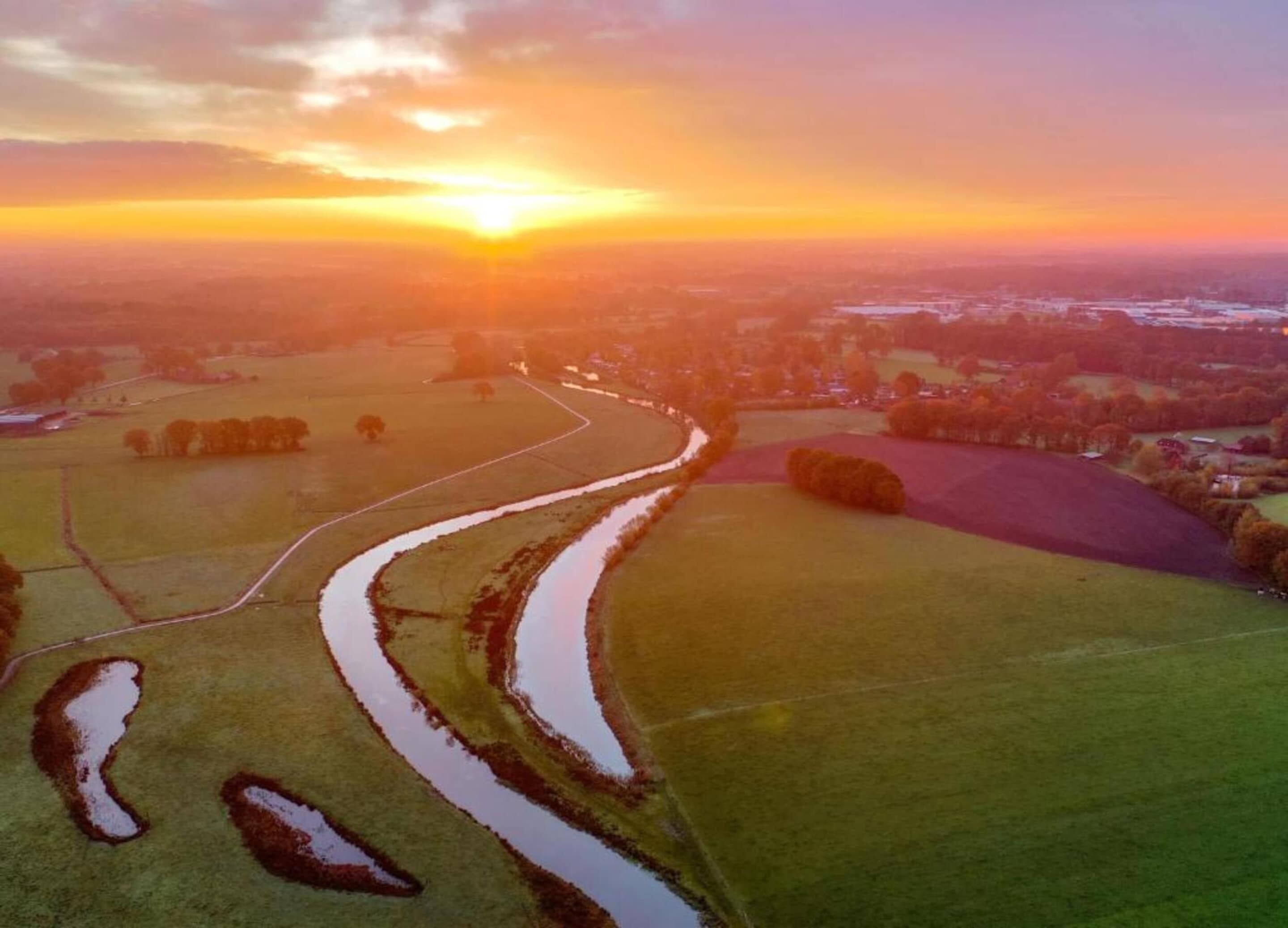 An aerial view of a rural landscape with fields, a winding water channel, and distant buildings.