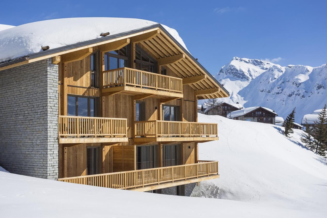 A multi-story wooden chalet with balconies, set against a snowy mountain backdrop.