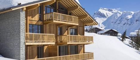 A multi-story wooden chalet with balconies, set against a snowy mountain backdrop.