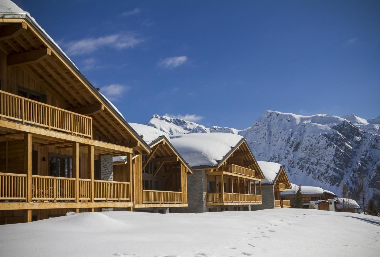 Una cabaña de madera con un balcón, frente a un paisaje montañoso cubierto de nieve.